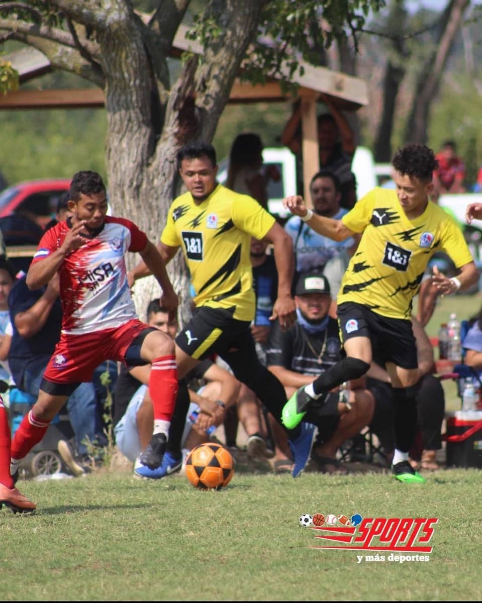 Tres jugadores disputan el balón en una cancha de pasto, dos de amarillo y uno de rojo, con aficionados en sillas plegables a la orilla.