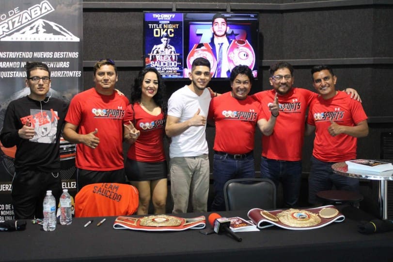 El equipo de Sports y Más Deportes, de playera roja, posa con el boxeador Alex Saucedo detrás de una mesa con dos cinturones de campeonato.